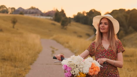 Middle-plan-a-young-sexy-blonde-on-a-bike-in-a-dress-and-flowers-in-a-basket-on-the-handlebar-goes-across-the-field-in-the-summer-walking-on-the-grass-in-slow-motion.
